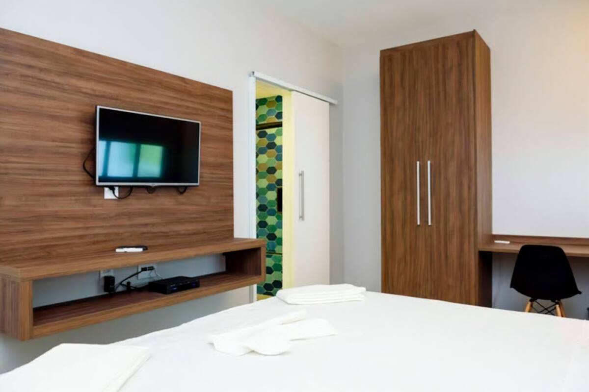Quarto de hotel moderno com um suporte de madeira para TV, cama com roupa de cama branca, escrivaninha com cadeira e guarda-roupa com duas portas.