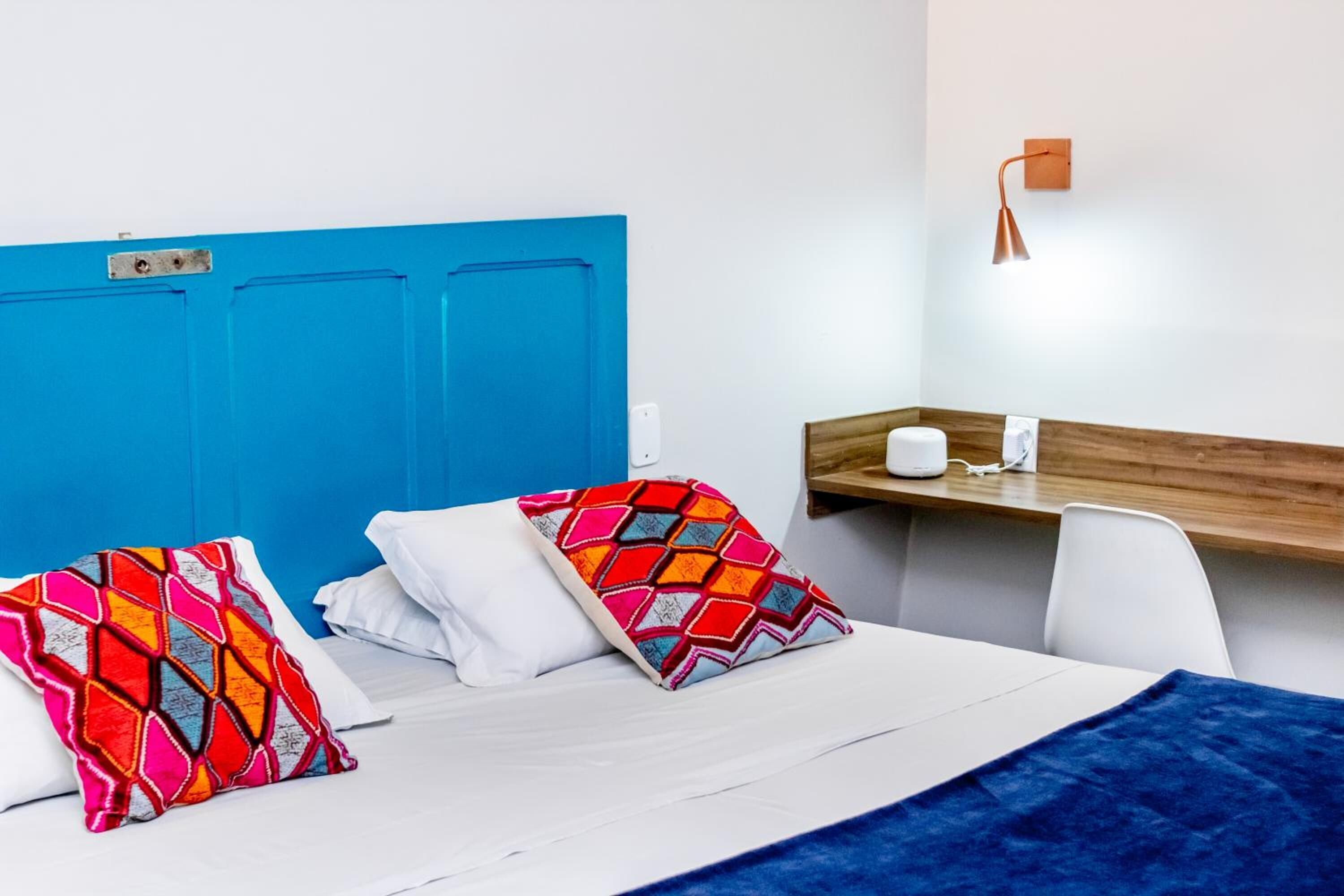 A hotel room with a blue headboard, two colorful pillows, a white bedspread, a blue blanket, a wooden side table with a lamp, and a white chair.