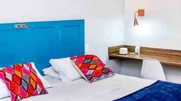 A hotel room with a blue headboard, two colorful pillows, a white bedspread, a blue blanket, a wooden side table with a lamp, and a white chair.