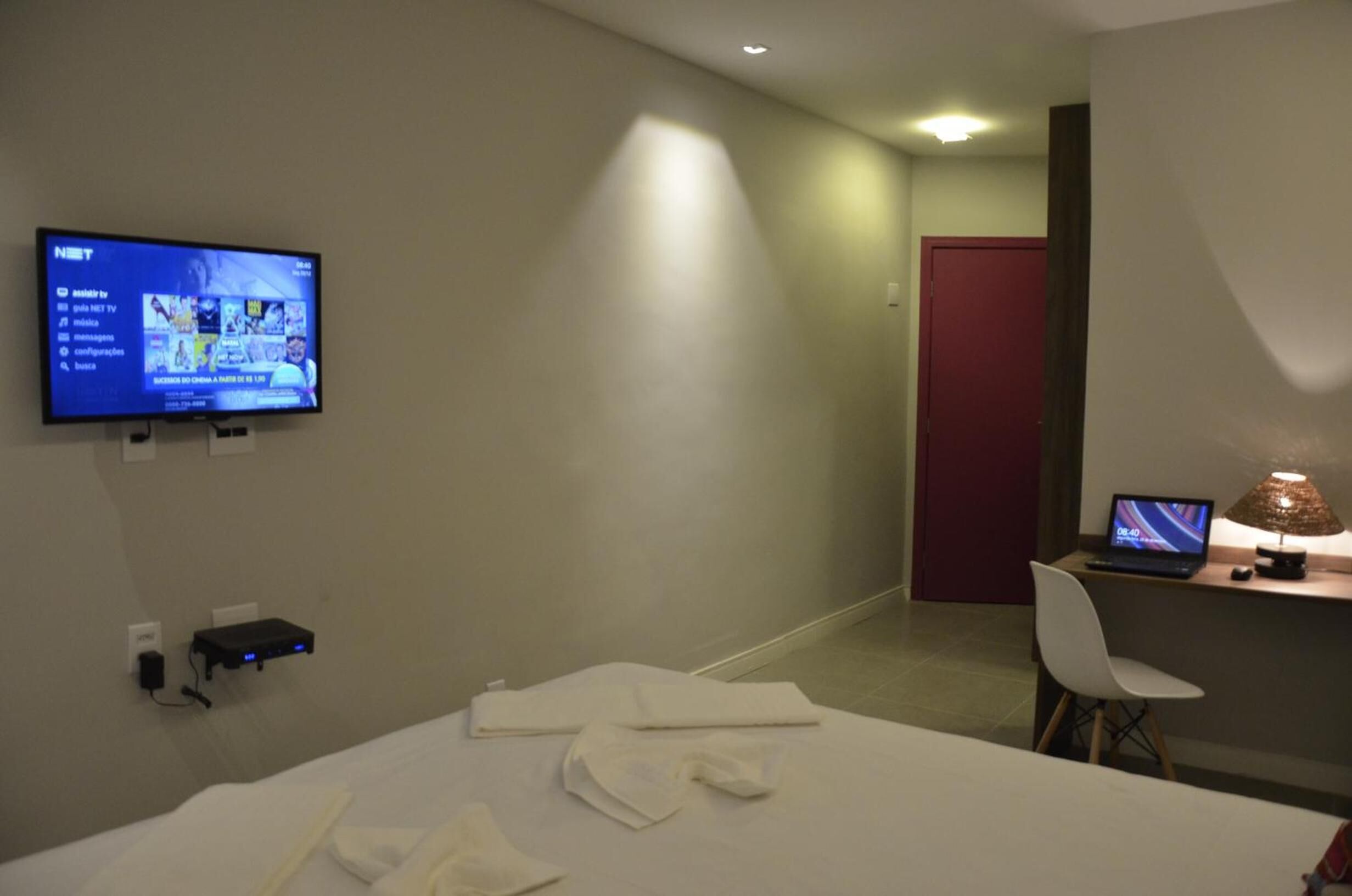 Habitación de hotel con una cama, un escritorio con una computadora portátil, un televisor montado en la pared y una puerta roja al fondo.