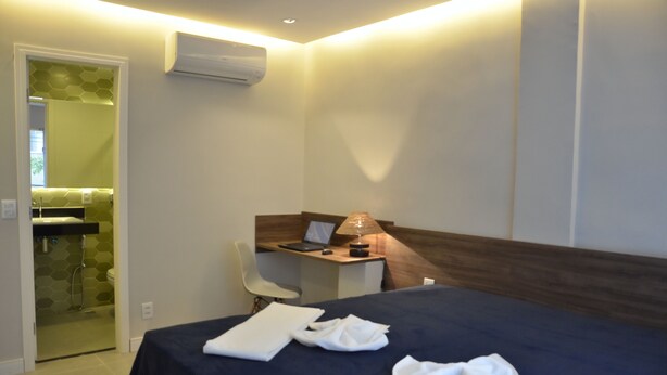 A hotel room with a bed, a desk with a laptop, a chair, and an air conditioning unit. - Icarai Suites (Niteroi)