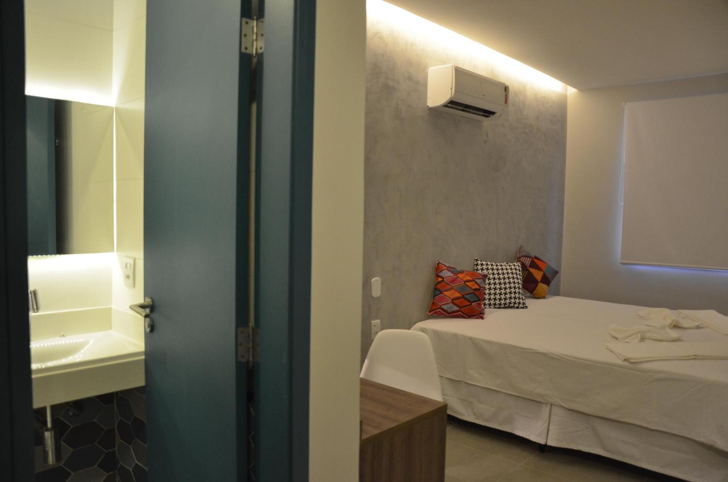 Un dormitorio con cama, mesita de noche, aire acondicionado de pared y baño con lavamanos y espejo.