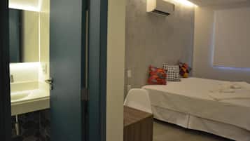 Un dormitorio con cama, mesita de noche, aire acondicionado de pared y baño con lavamanos y espejo.