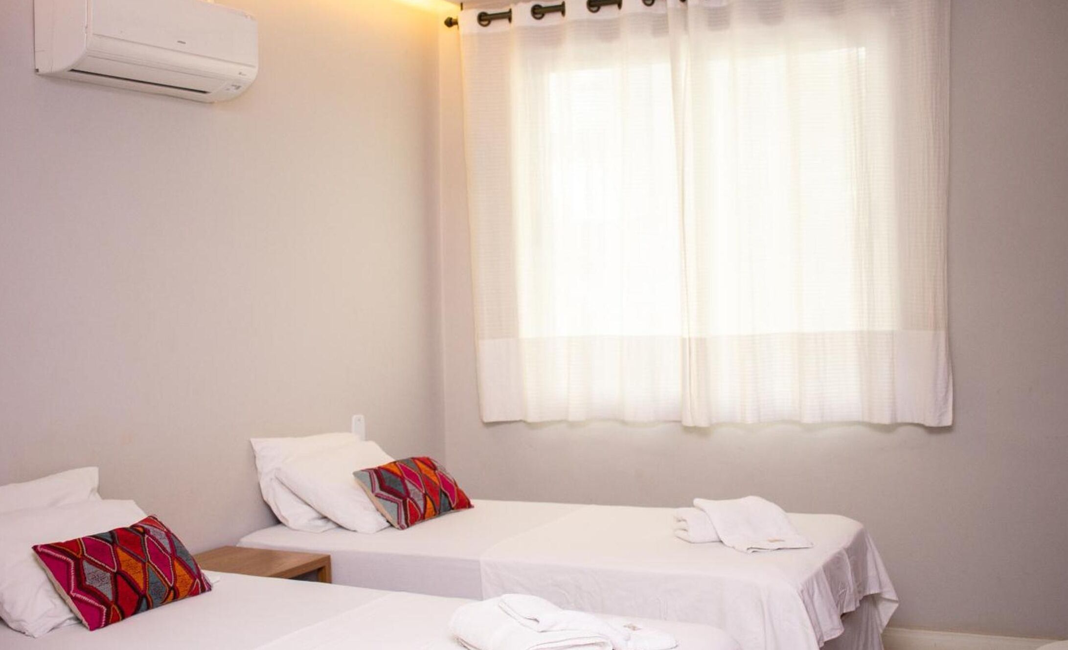 Una habitación de hotel con dos camas individuales, una ventana con cortinas traslúcidas, un aire acondicionado de pared y una mesita de noche con lámpara.
