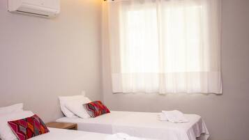 Una habitación de hotel con dos camas individuales, una ventana con cortinas traslúcidas, un aire acondicionado de pared y una mesita de noche con lámpara.