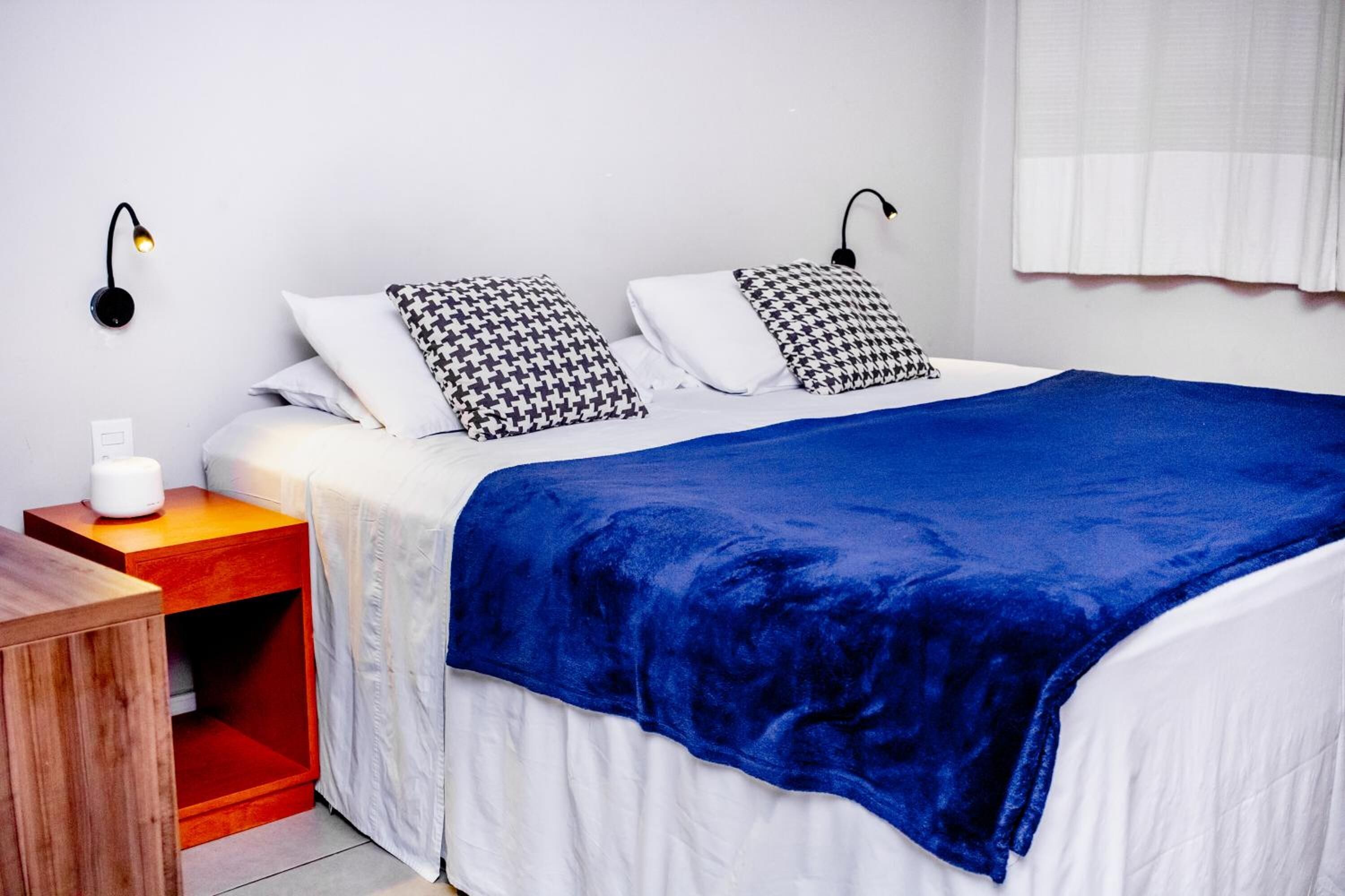 A neatly made bed with a blue blanket, two patterned pillows, a wooden nightstand with a white object on it, and two wall-mounted lamps.