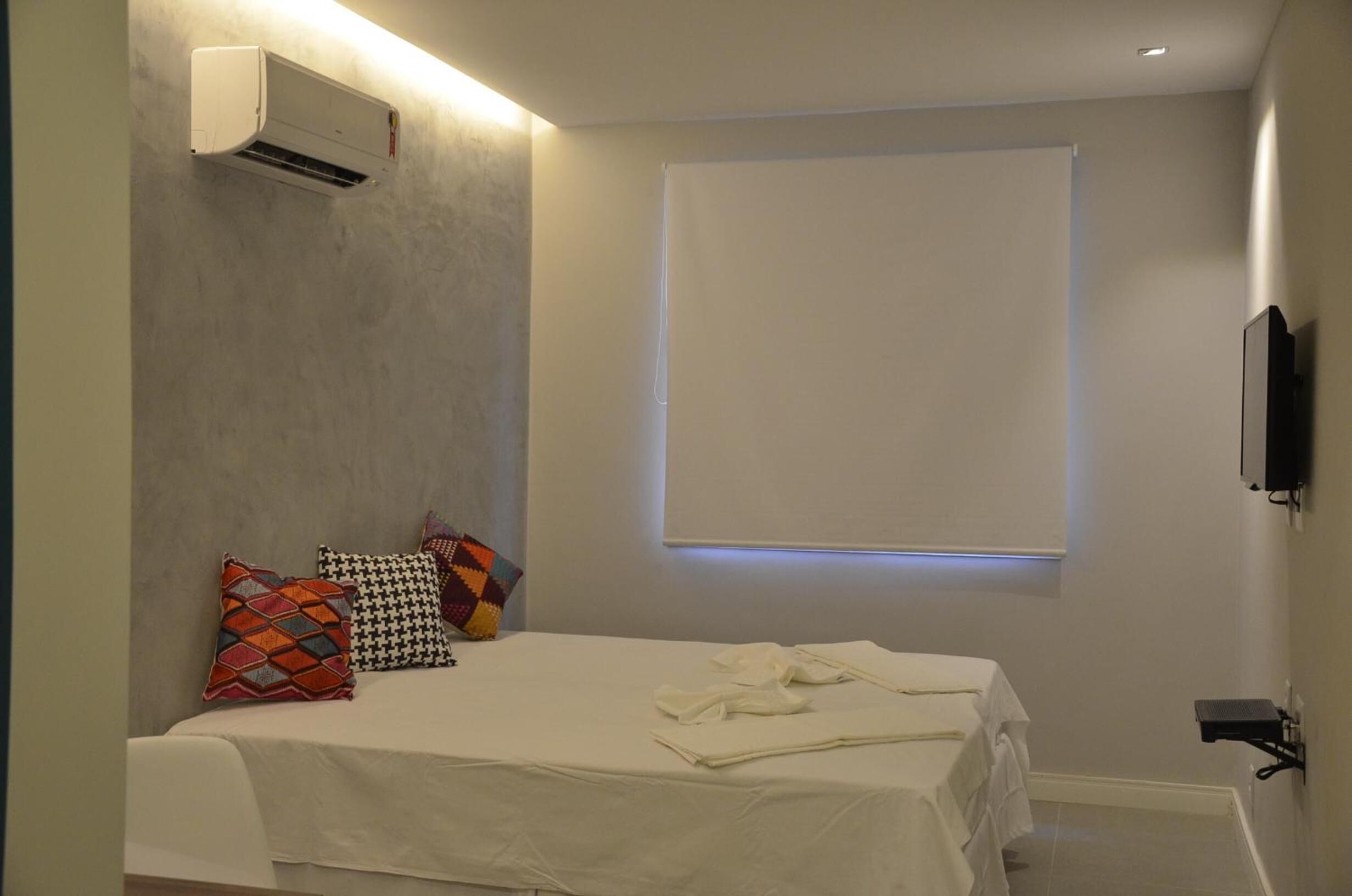 Una camera da letto con un letto, cuscini, condizionatore d'aria, televisore e una finestra con persiane.