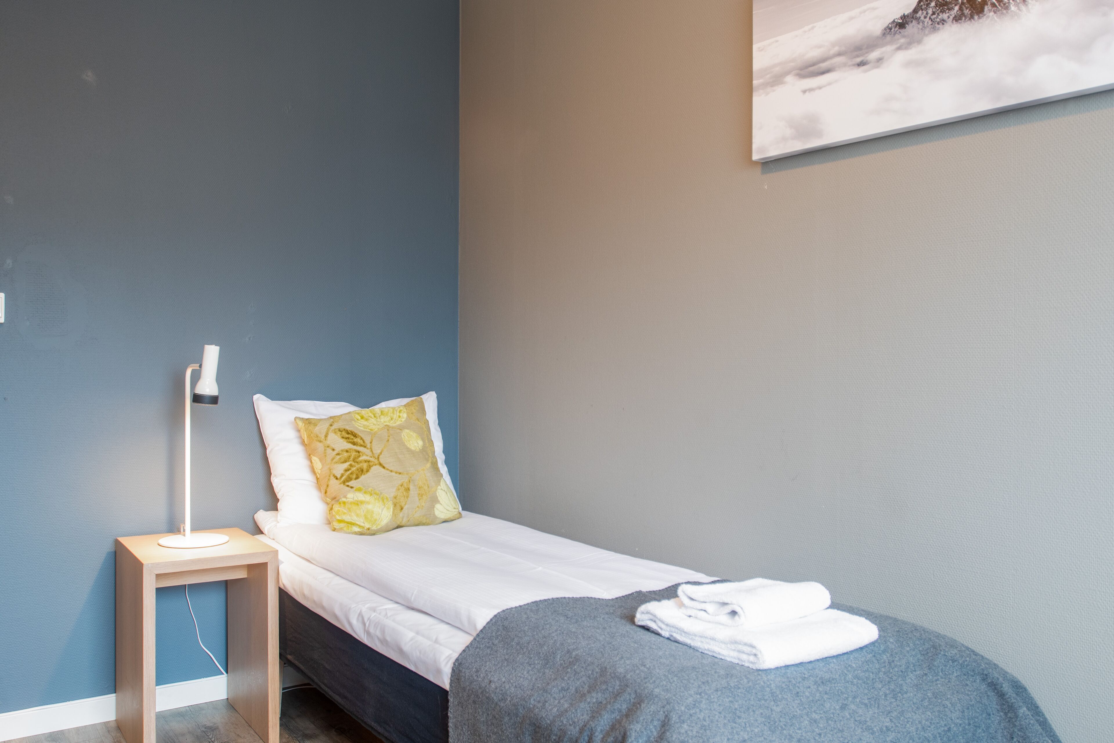 A neatly made bed with a white sheet and a blue blanket, a bedside lamp, and a pillow with a yellow pattern.