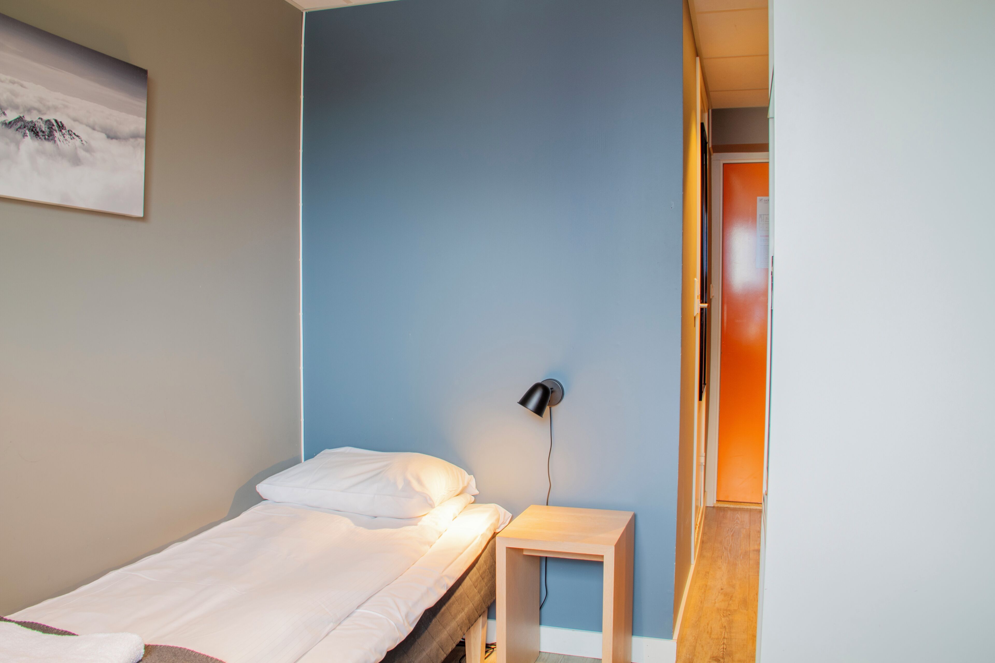 Une chambre à coucher comprenant un lit, une table de chevet, une lampe fixée au mur et une porte donnant sur une autre pièce.
