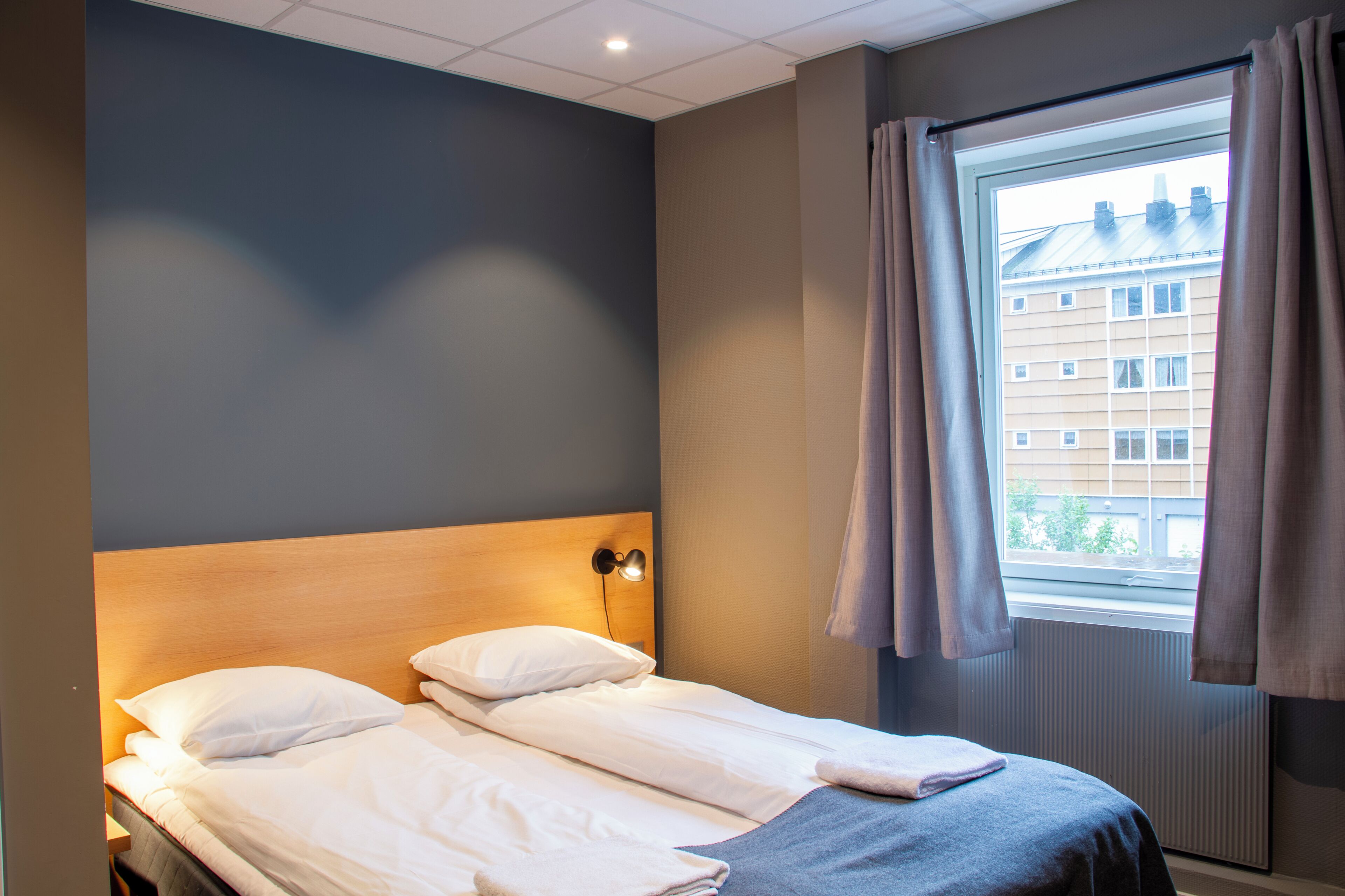 A hotel room with a bed, a headboard, a window with curtains, and a wall-mounted light.