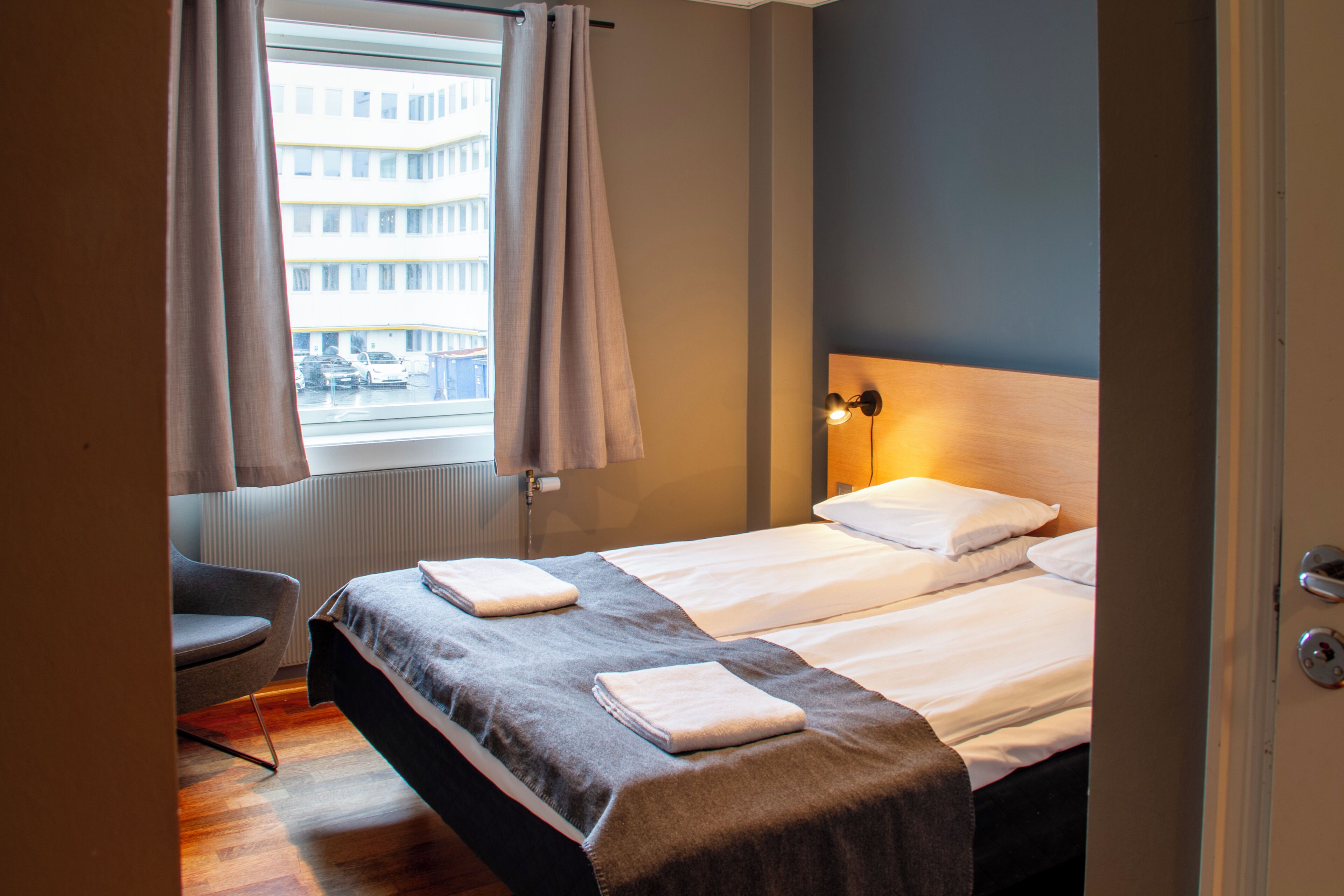 A hotel room with a bed, a chair, a window with curtains, a radiator, and a view of a building outside.