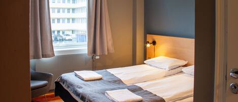 A hotel room with a bed, a chair, a window with curtains, a radiator, and a view of a building outside.