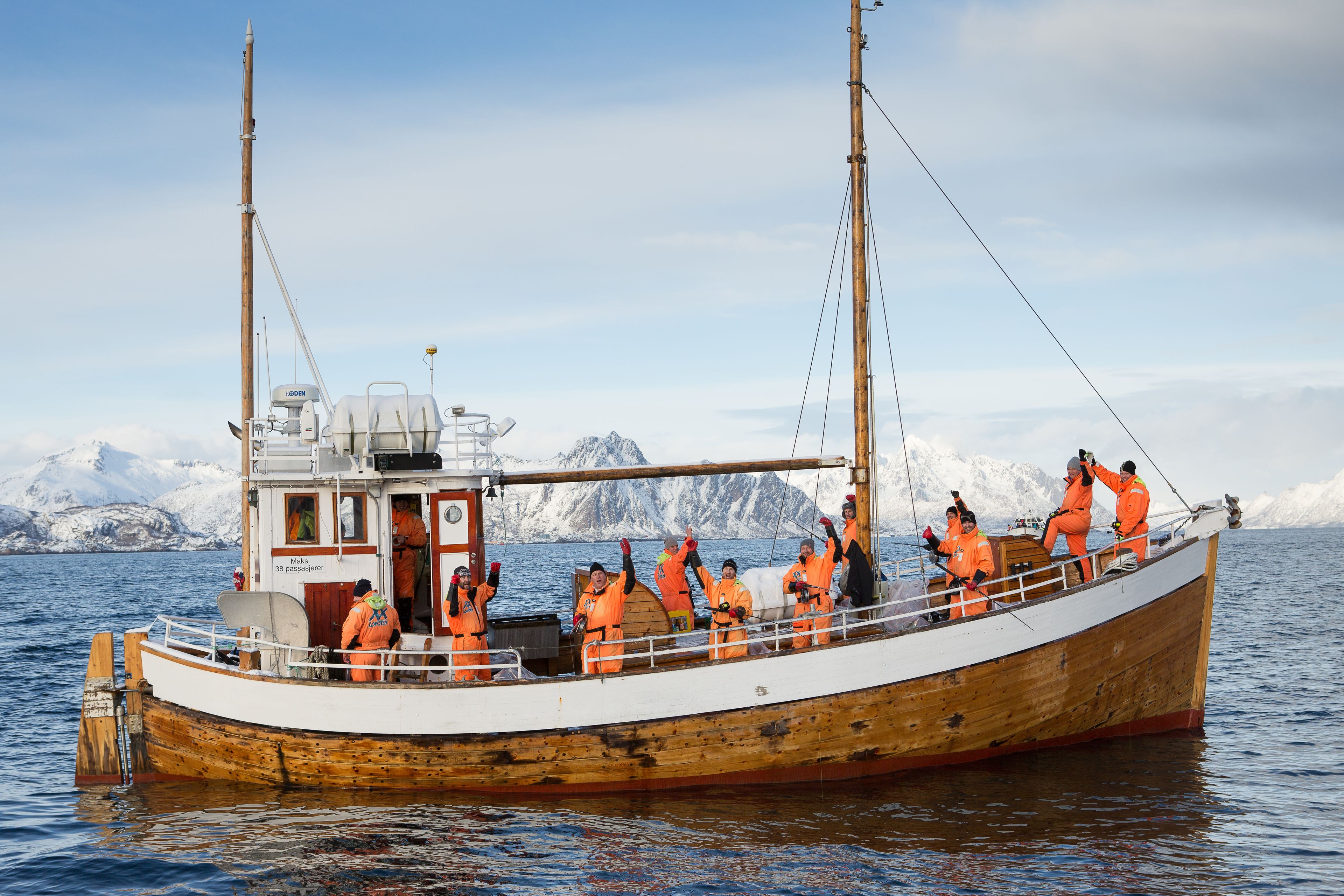 A wooden boat with passengers wearing orange life jackets, anchored in a body of water with a mountainous landscape in the background.