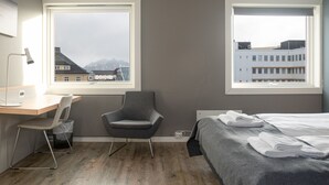A hotel room with a wooden floor, a desk with a chair, a bed with towels, and two windows offering views of buildings and mountains.