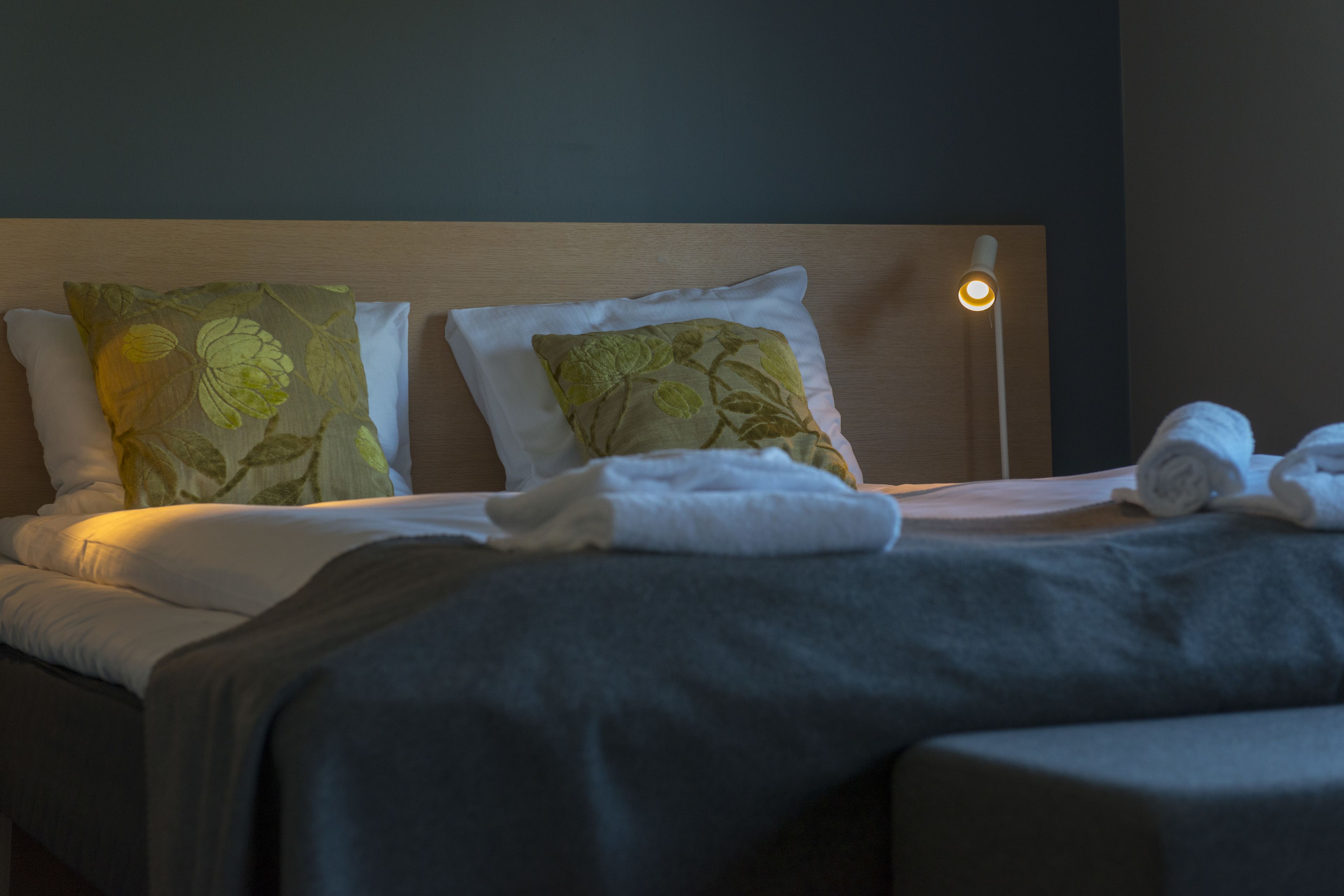 A neatly made bed with white linens and two decorative pillows, a bedside lamp, and a rolled towel on the bed.
