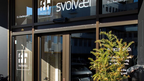 Una entrada de hotel moderna con puertas de vidrio, una jardinera de madera con una planta, y el nombre del hotel 'Svolvaer' exhibido en las ventanas.