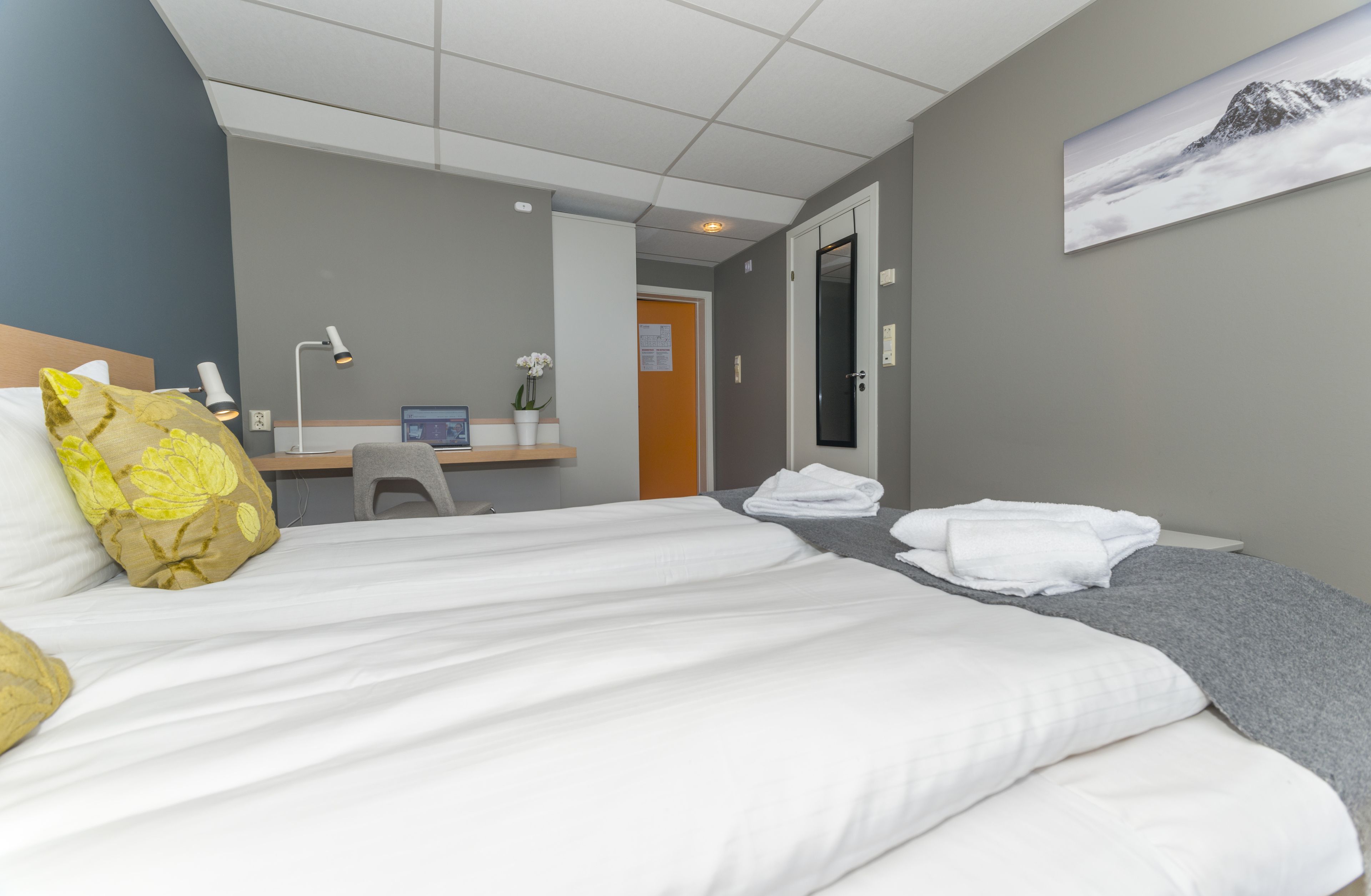 A hotel room with a bed, a desk, and a chair. There is a door leading to another room and a painting on the wall.