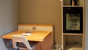 Una habitación pequeña con un escritorio de madera, una silla, un bote de basura y un estante con una tetera y una taza.