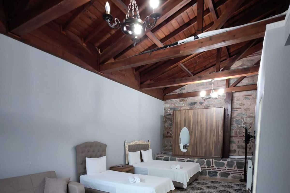 A spacious room with a high ceiling and exposed wooden beams. It features two beds with white linens, a sofa, a wooden coffee table, and a patterned rug.