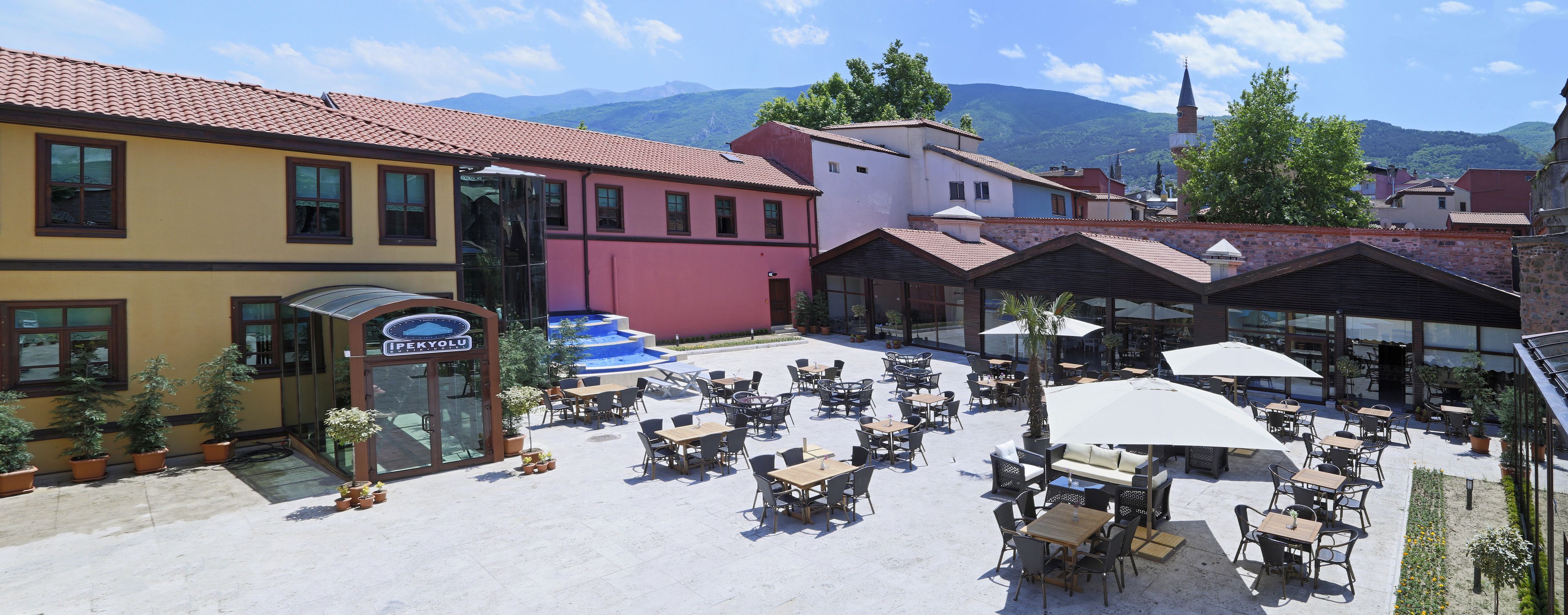 A courtyard with outdoor seating, a building with a sign reading "PEKYOLU," and a mountain backdrop.