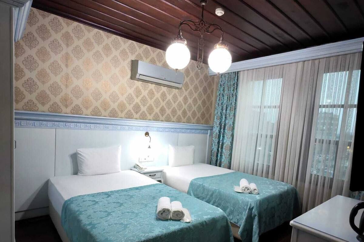 A hotel room with two beds, a wooden ceiling, an air conditioning unit, a patterned rug, and a small desk with a kettle.