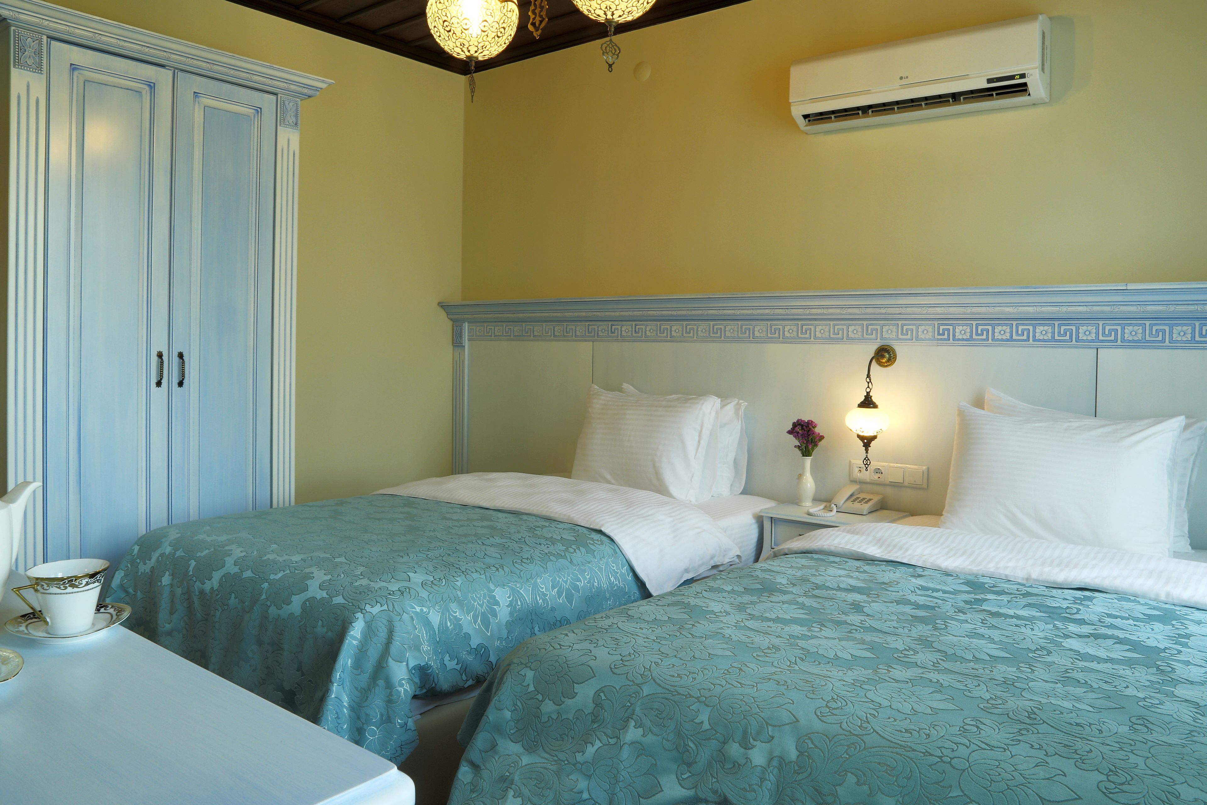 A hotel room with two beds, a wardrobe, a bedside table with a lamp, and an air conditioning unit.