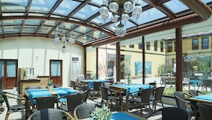 Un espacio comedor al aire libre con techado, mesas y sillas, techo de cristal y vista a edificios.
