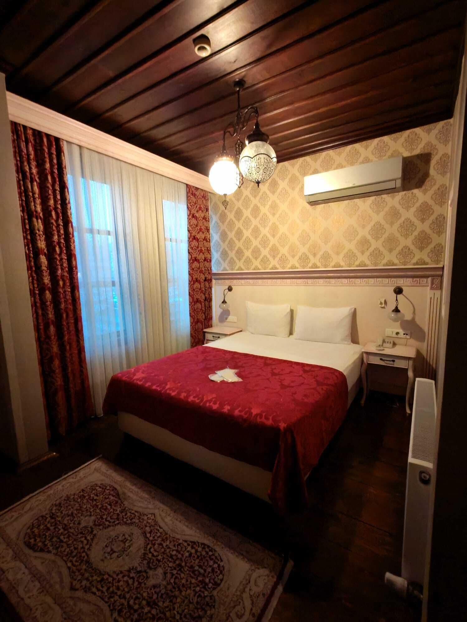 Habitación de hotel con techo de madera, cama con colcha roja, alfombra estampada, mesita de noche con lámpara y unidad de aire acondicionado en la pared.