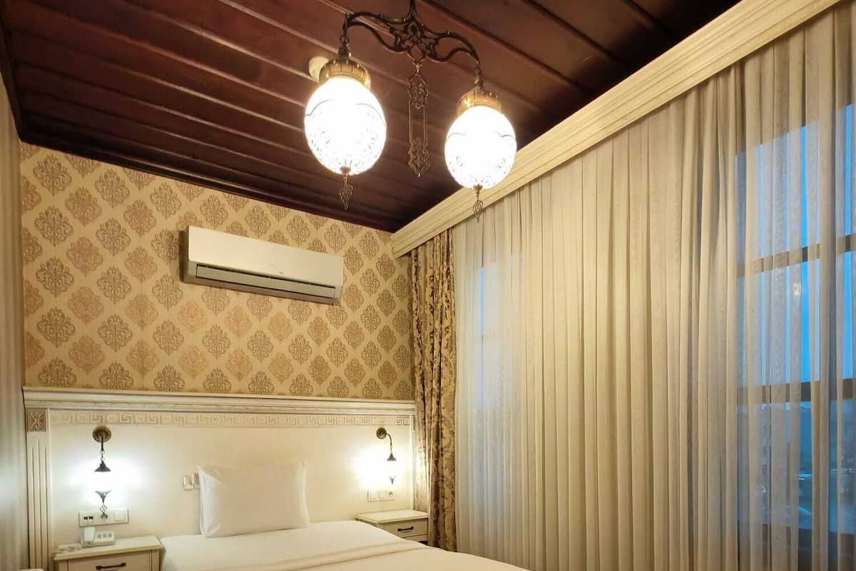 A hotel room with a large bed, two bedside lamps, an air conditioner, a patterned rug, and a wooden ceiling.
