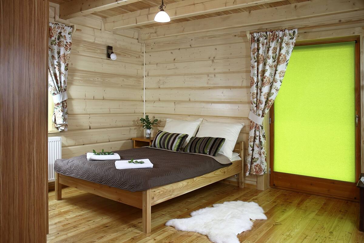 A wooden cabin bedroom with a bed, curtains, a nightstand, and a small plant.