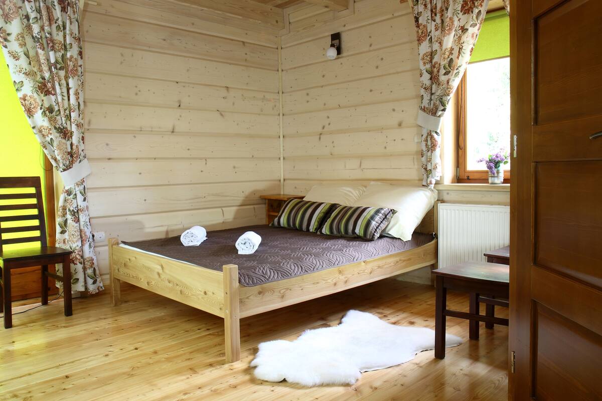 A wooden cabin room with a bed, a chair, a table, and a fur rug.