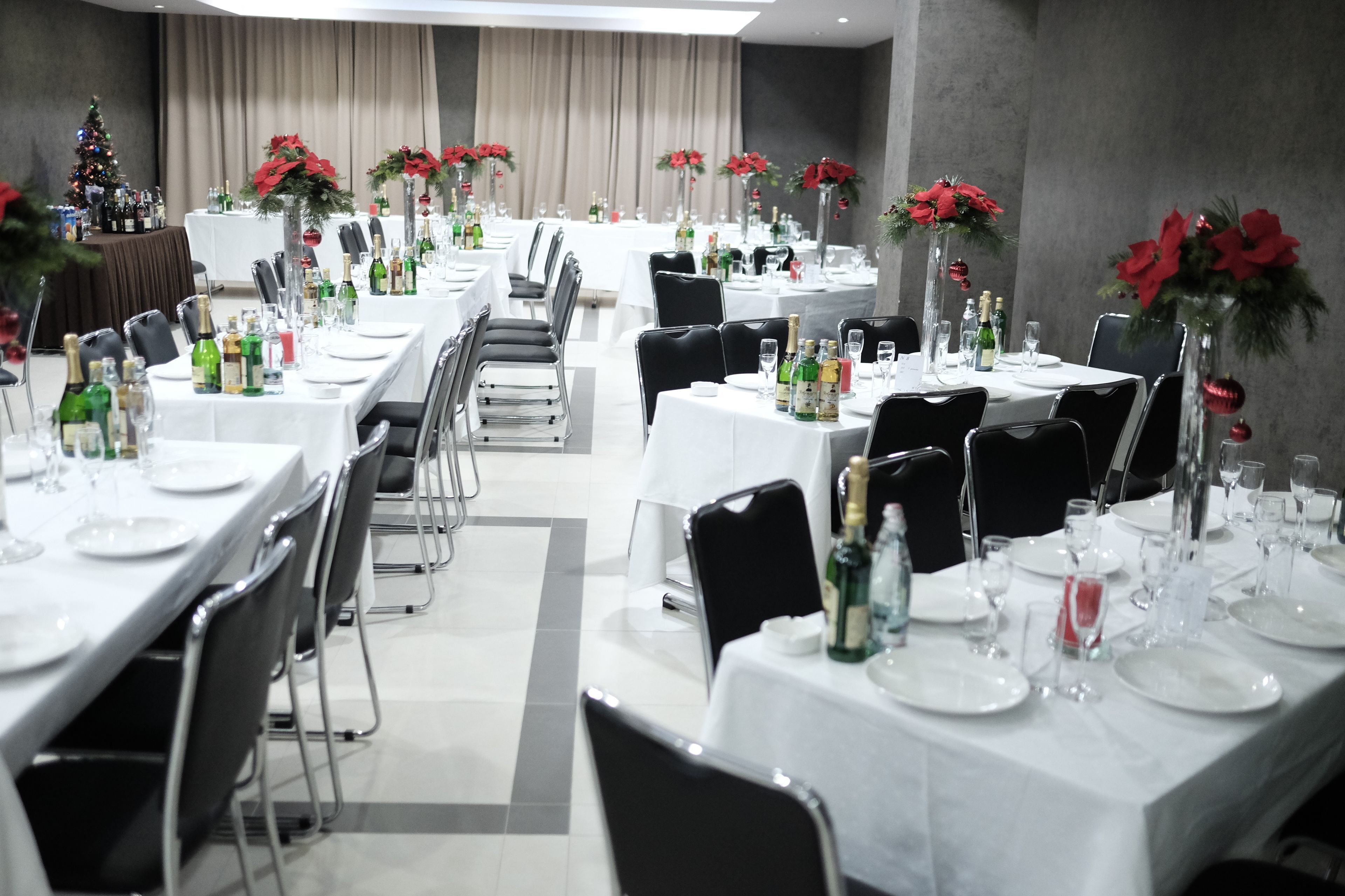 A hotel dining area with multiple tables set for a formal event, featuring white tablecloths, black chairs, and red poinsettia centerpieces.