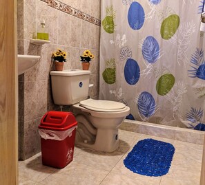 A bathroom with a toilet, a red trash bin, a blue pebble rug, and a shower curtain with a leaf pattern.