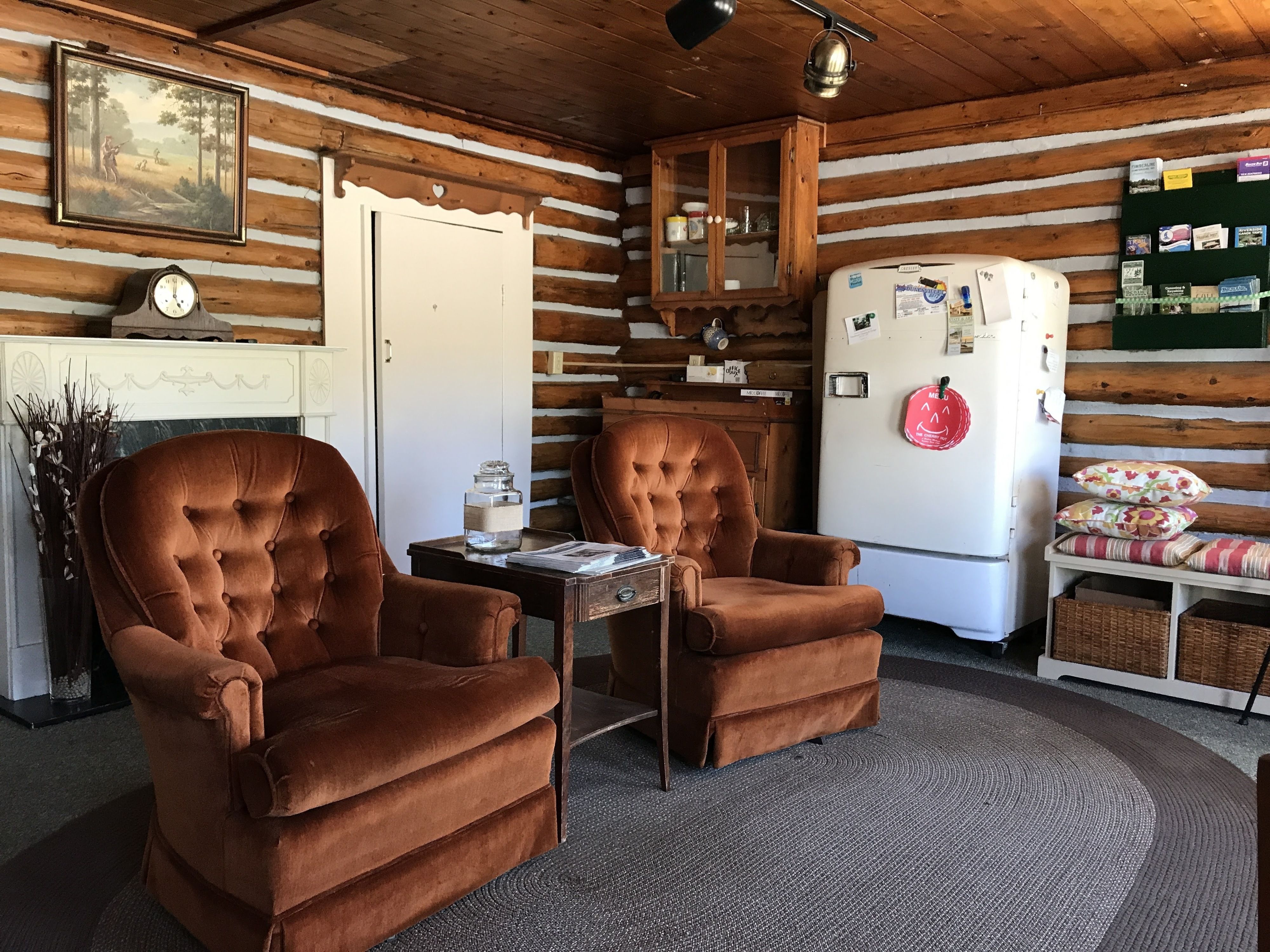 A cozy log cabin interior with two brown tufted chairs, a white refrigerator, a wooden cabinet, and a clock on the wall.
