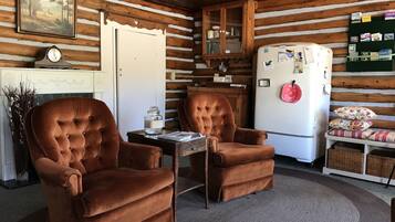 A cozy log cabin interior with two brown tufted chairs, a white refrigerator, a wooden cabinet, and a clock on the wall.