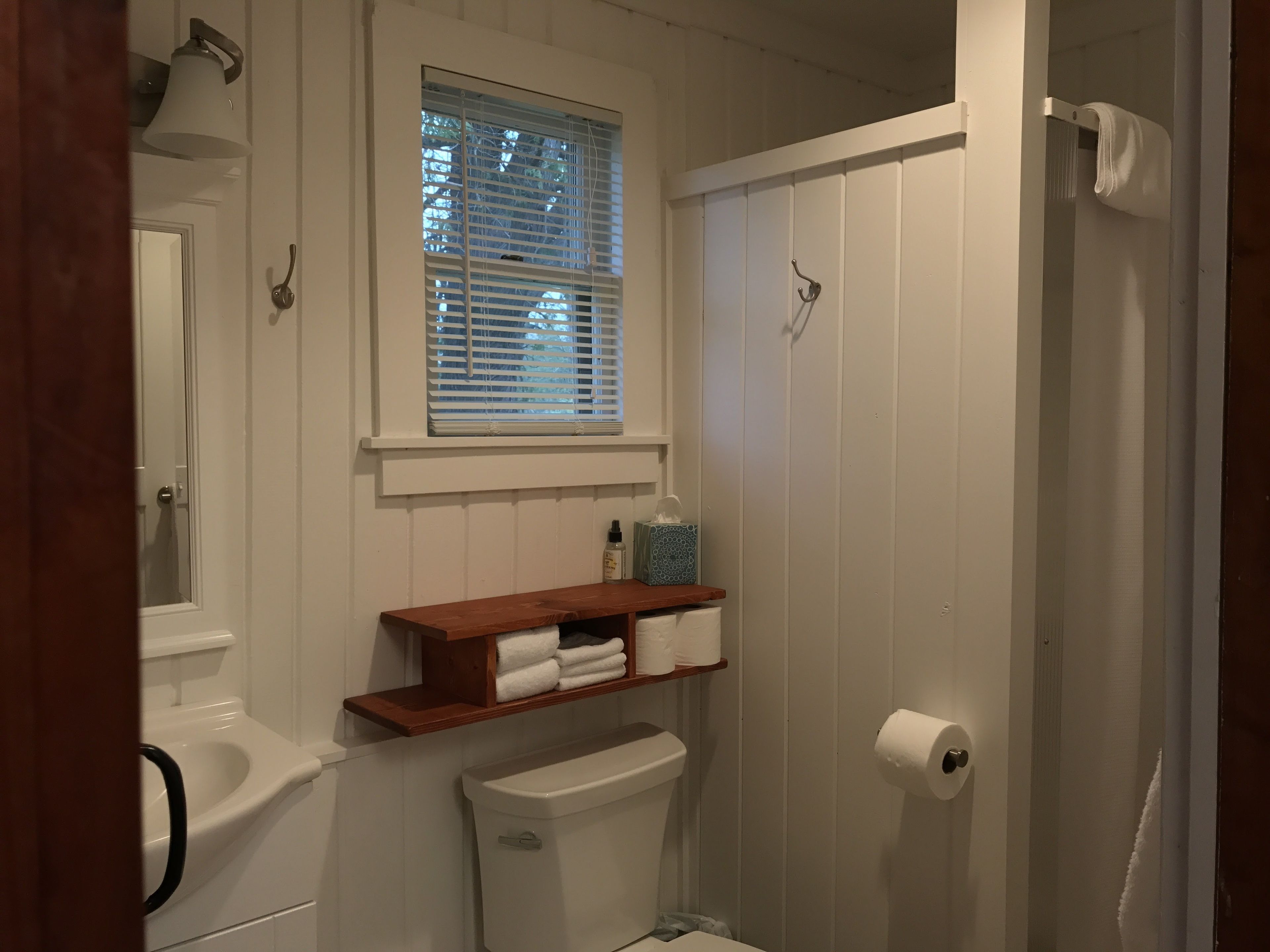 A bathroom with a toilet, a sink, a window with blinds, a towel rack, and a shelf with towels.