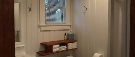 A bathroom with a toilet, a sink, a window with blinds, a towel rack, and a shelf with towels.