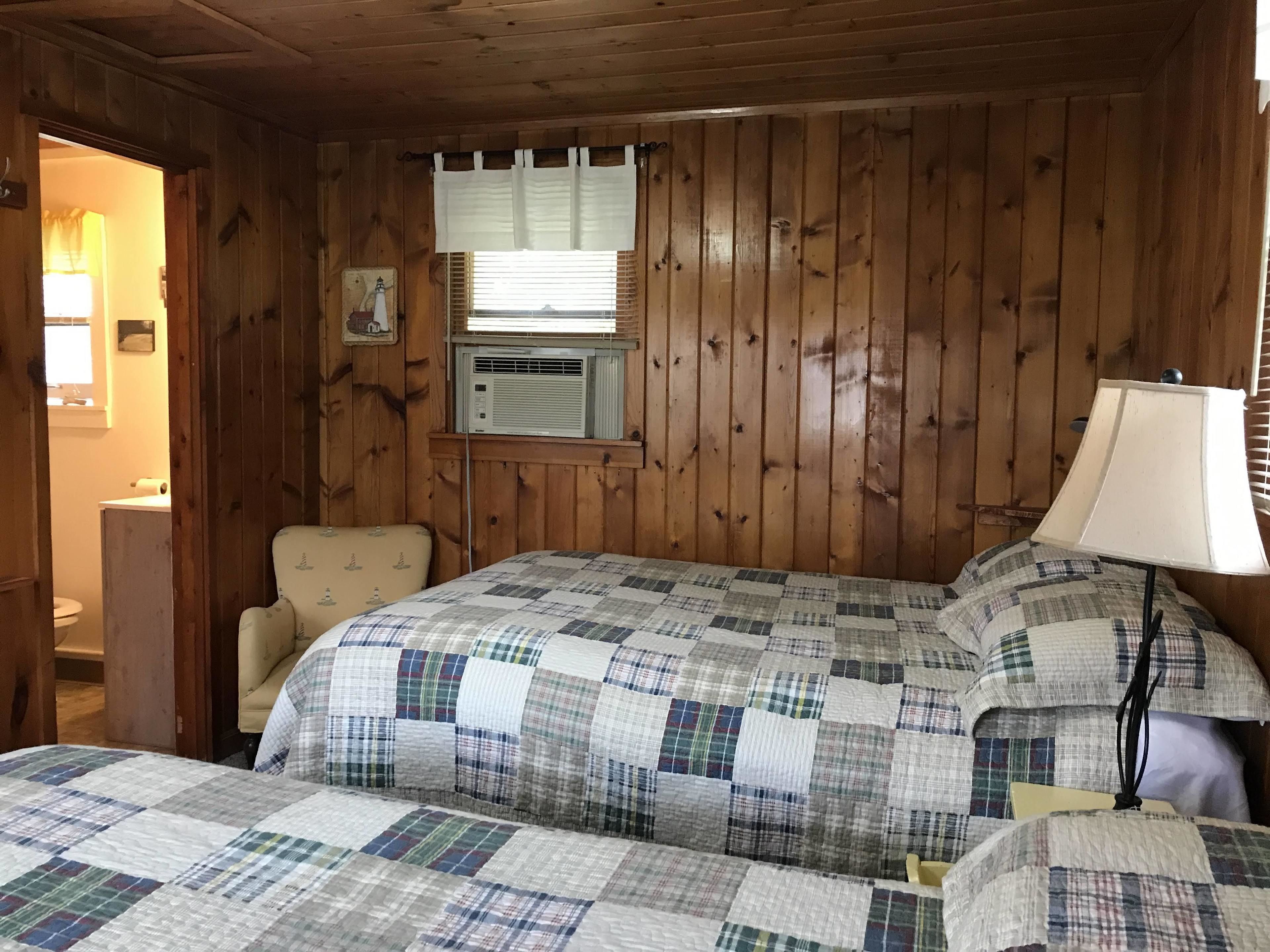 A room with wooden paneling, a bed with a checkered quilt, a bedside lamp, and a window with blinds.