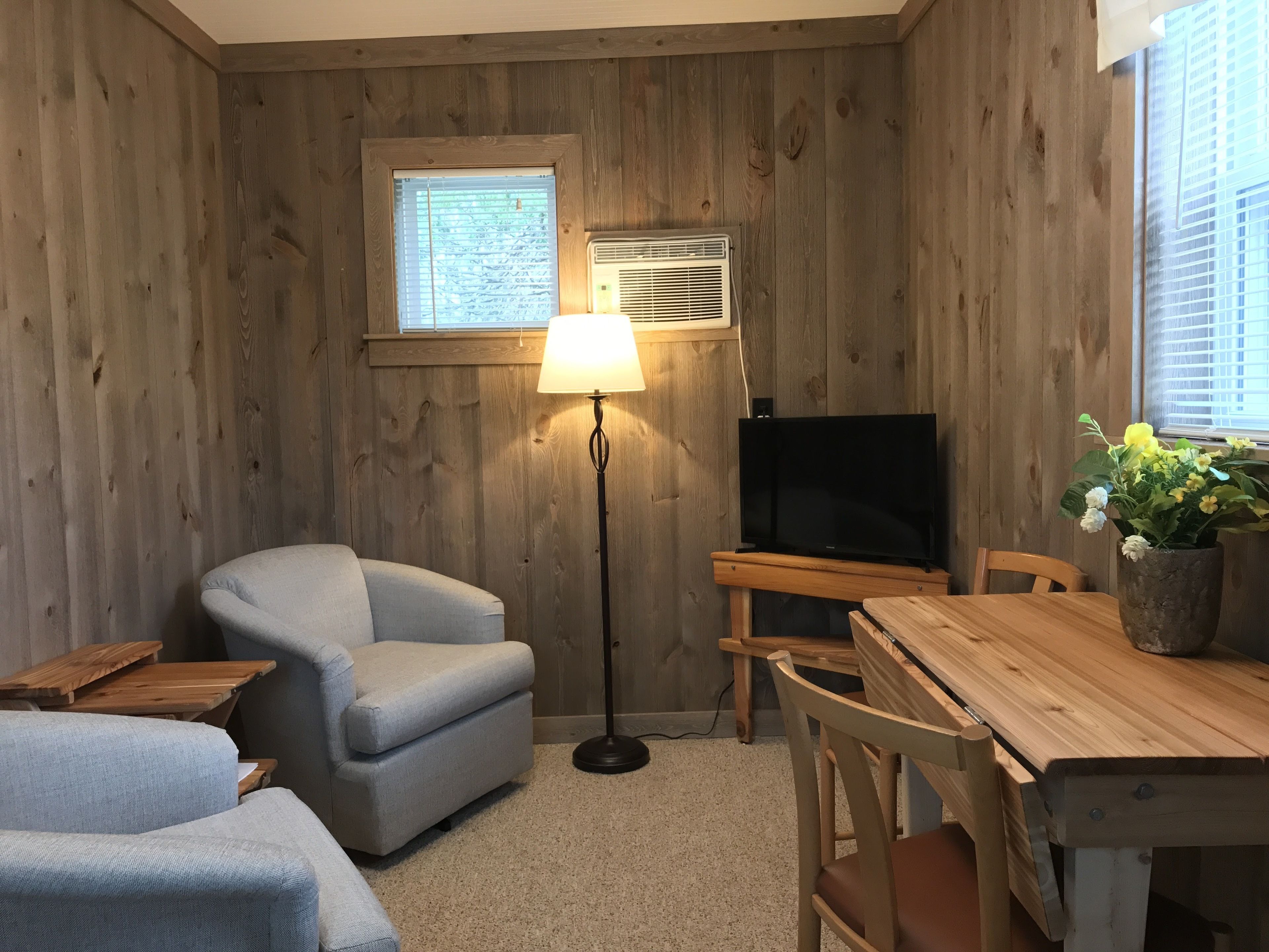 A cozy room with wooden walls, a comfortable armchair, a small table, a TV, and a window with blinds.