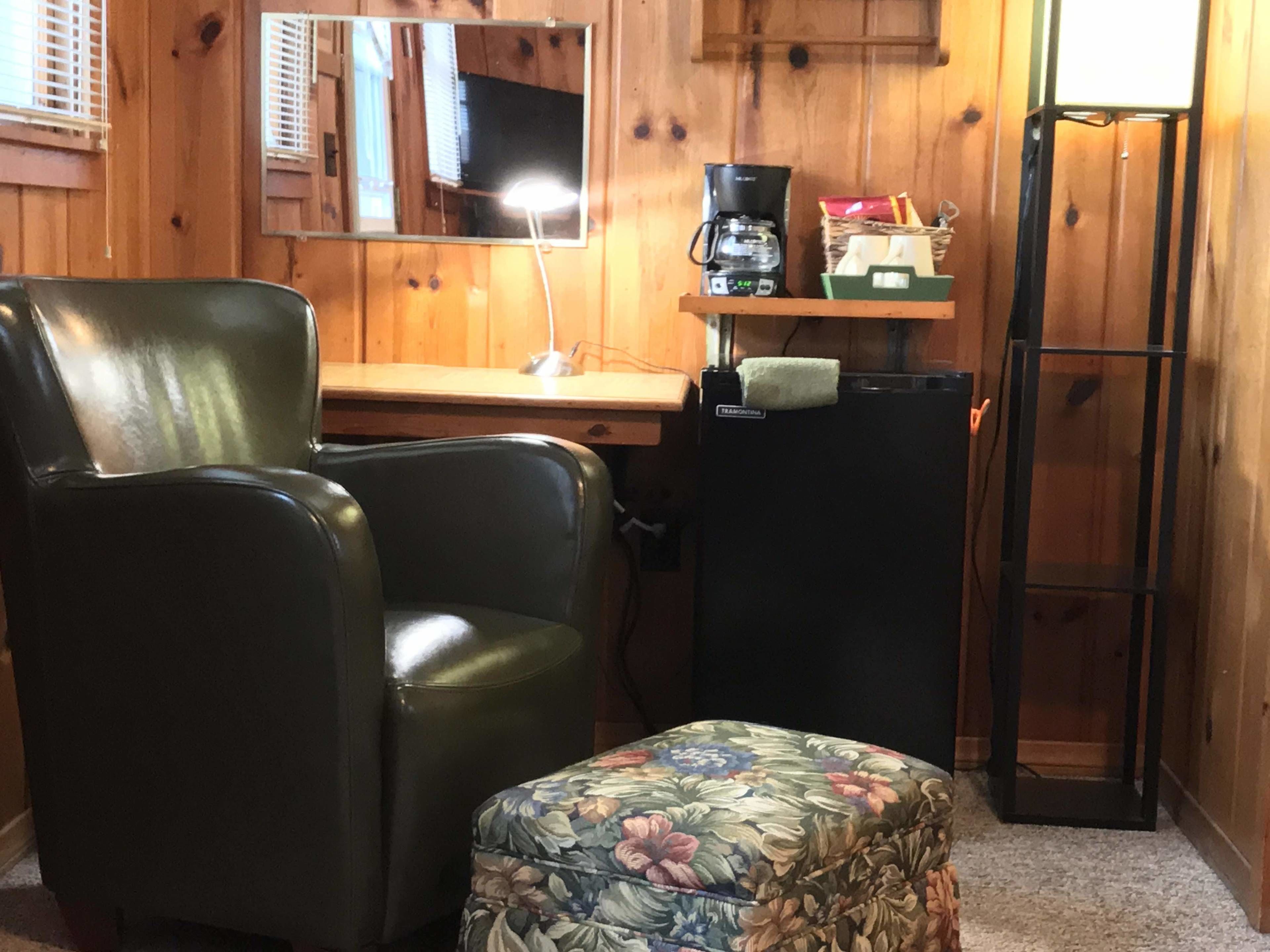 A room with a leather chair, a small floral ottoman, a wooden desk with a coffee maker, and a mirror on the wall.