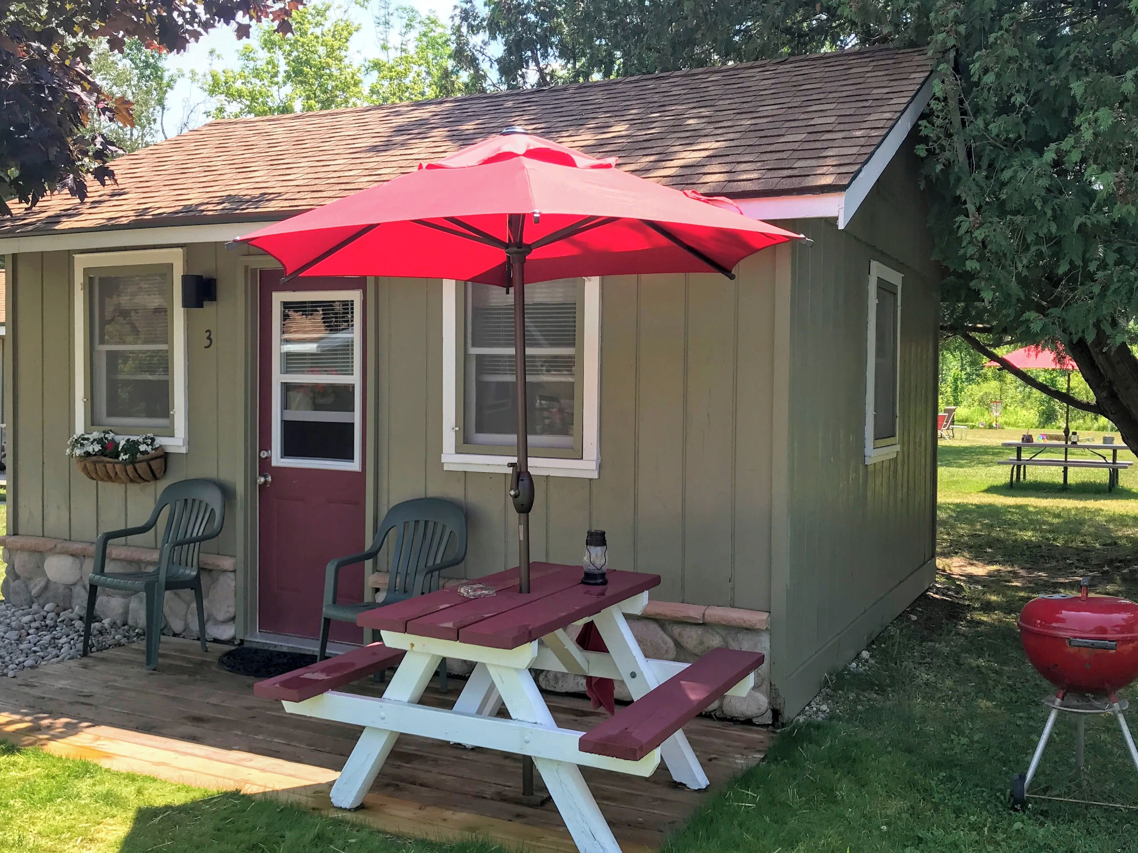A small cabin with a picnic table and two chairs, a red umbrella, and a barbecue grill.