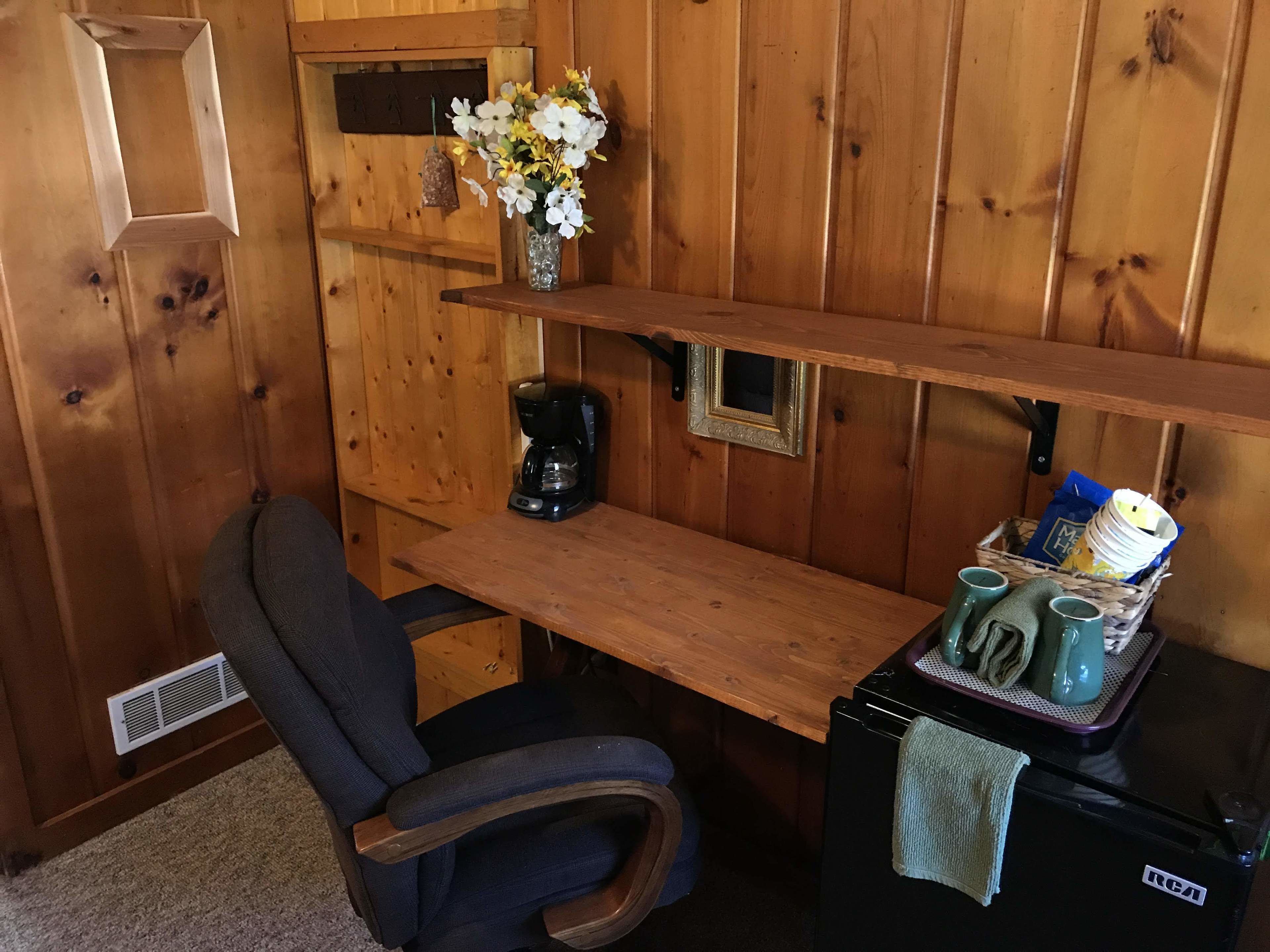 A wooden cabin interior with a wooden chair, a small table, a coffee maker, a vase with flowers, a framed picture, and a refrigerator with a towel on it.