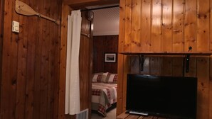 A wooden-paneled room with a bed, a television on a wooden stand, and a curtain partially drawn. - Cedar Ridge Cabins (Honor)