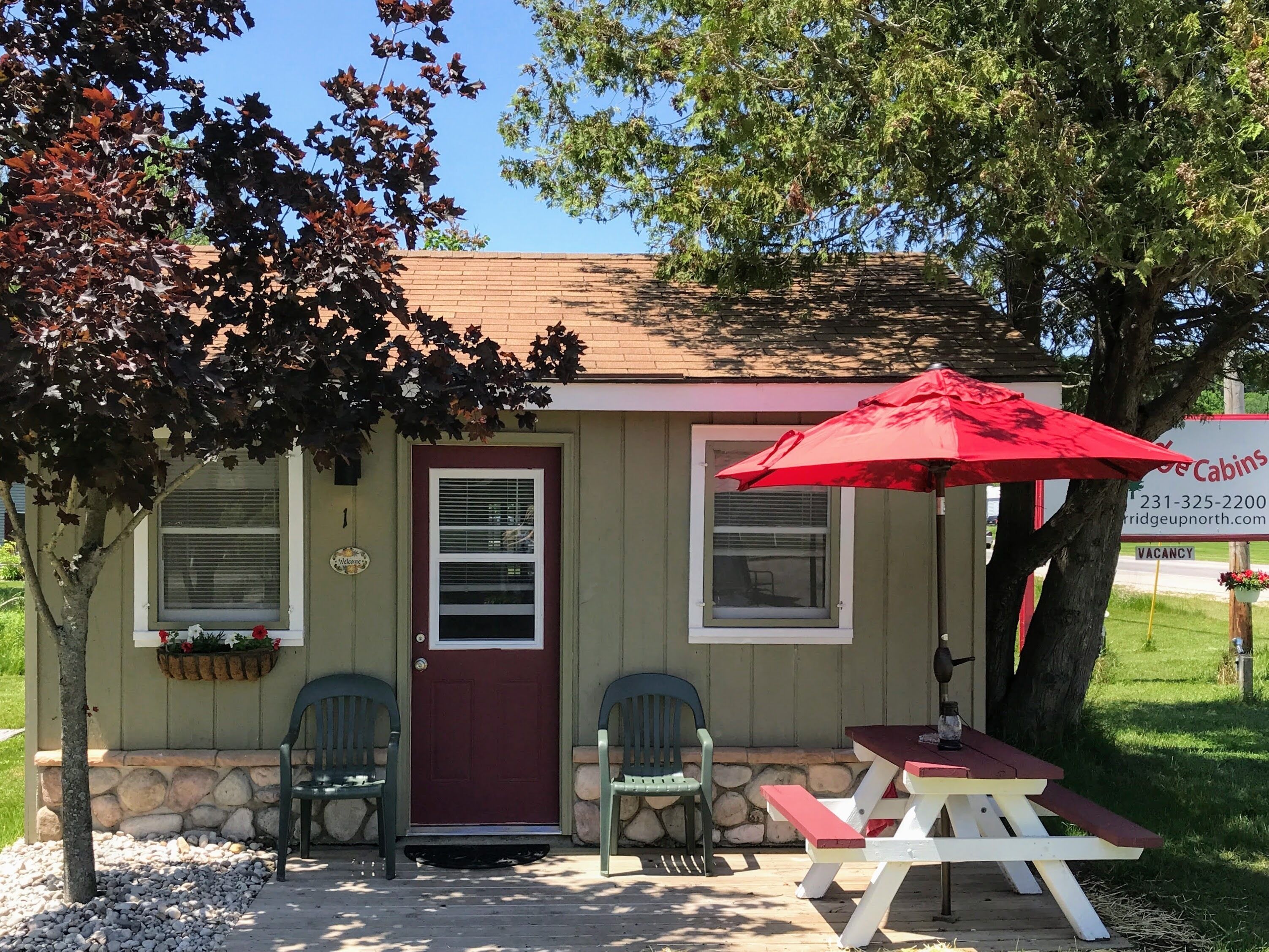 A small building with a red door, two windows, and a picnic table with a red umbrella. There are two chairs and a stone wall.