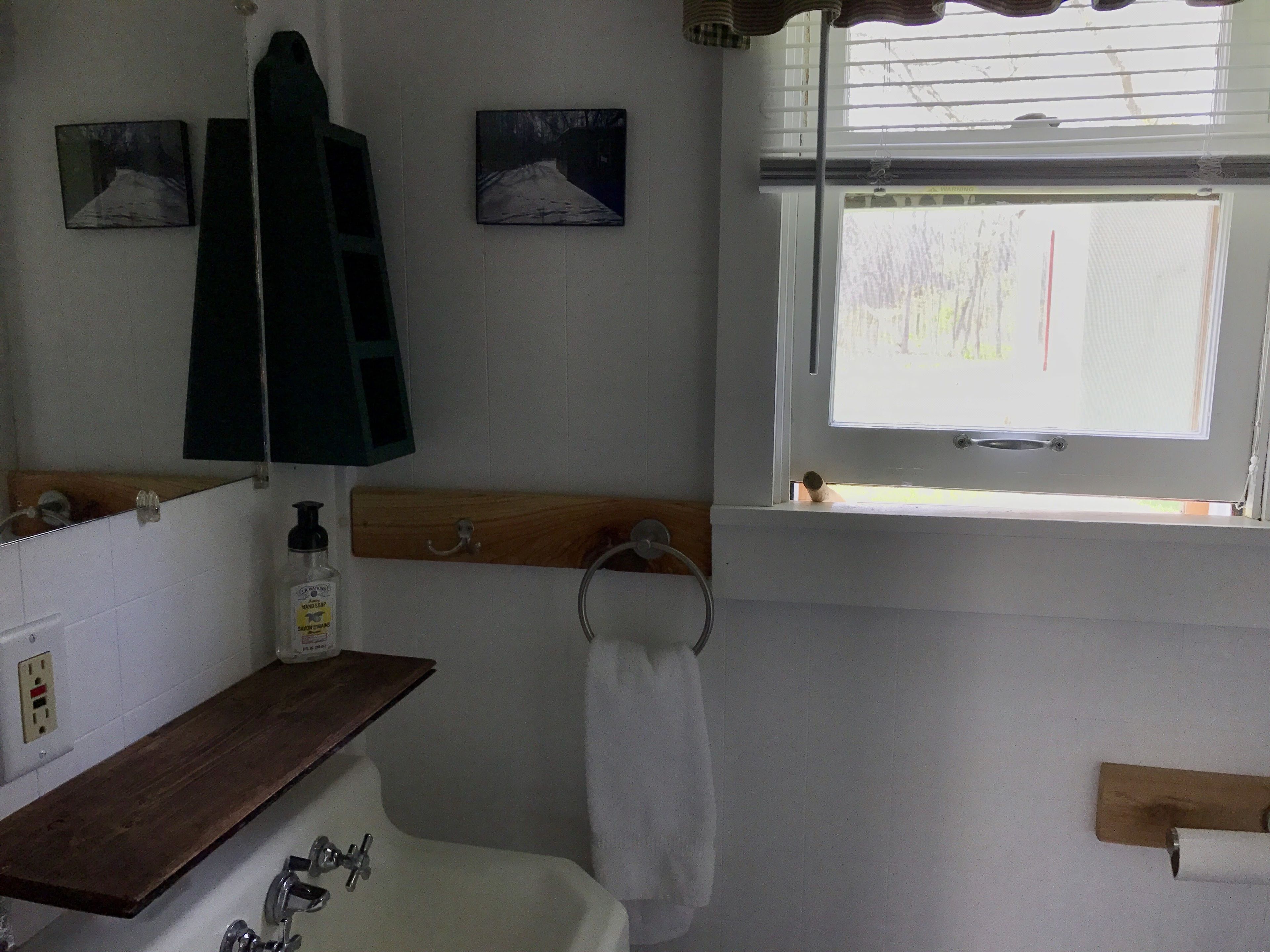 Un baño con un lavamanos blanco, una estantería de madera, un perchero para toallas y una ventana con persianas.