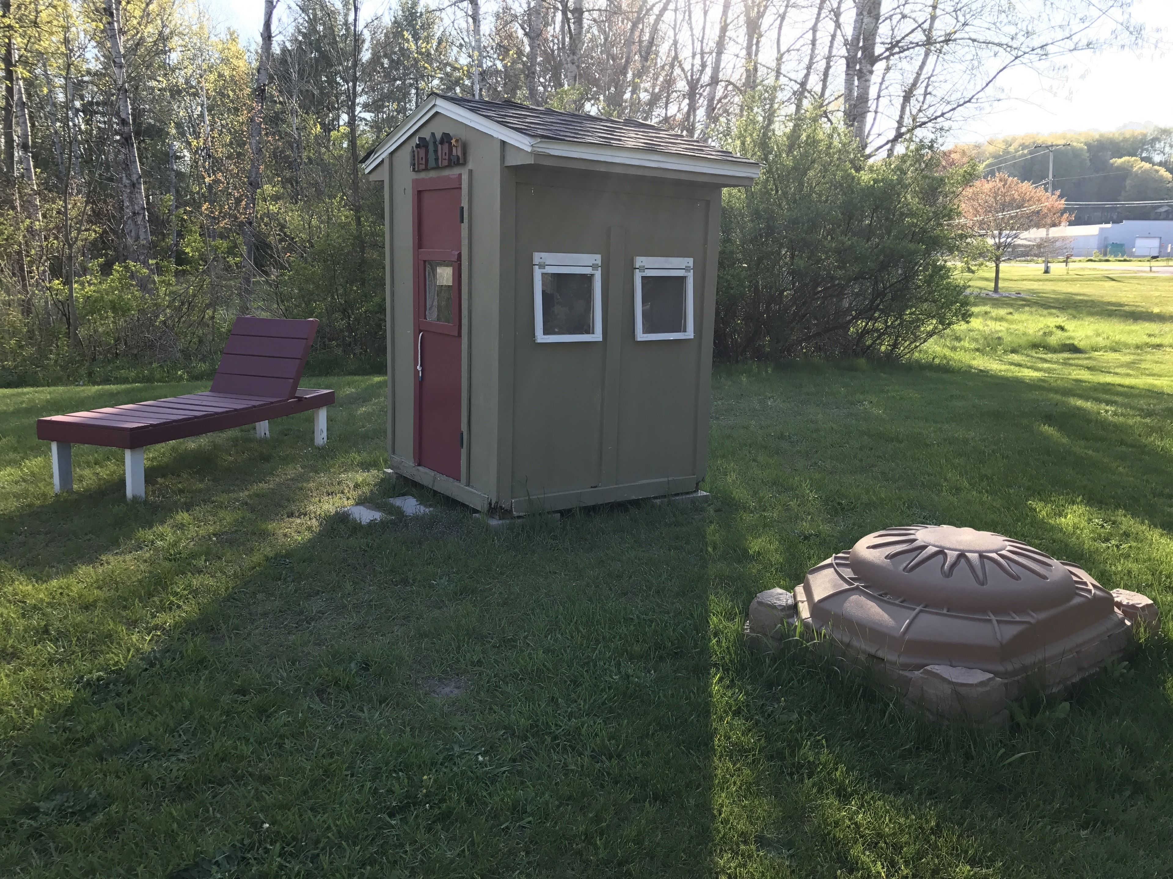 Un petit abri de jardin avec une porte rouge et deux fenêtres, un fauteuil de jardin rouge et une structure décorative en pierre, le tout situé dans un espace verdoyant.