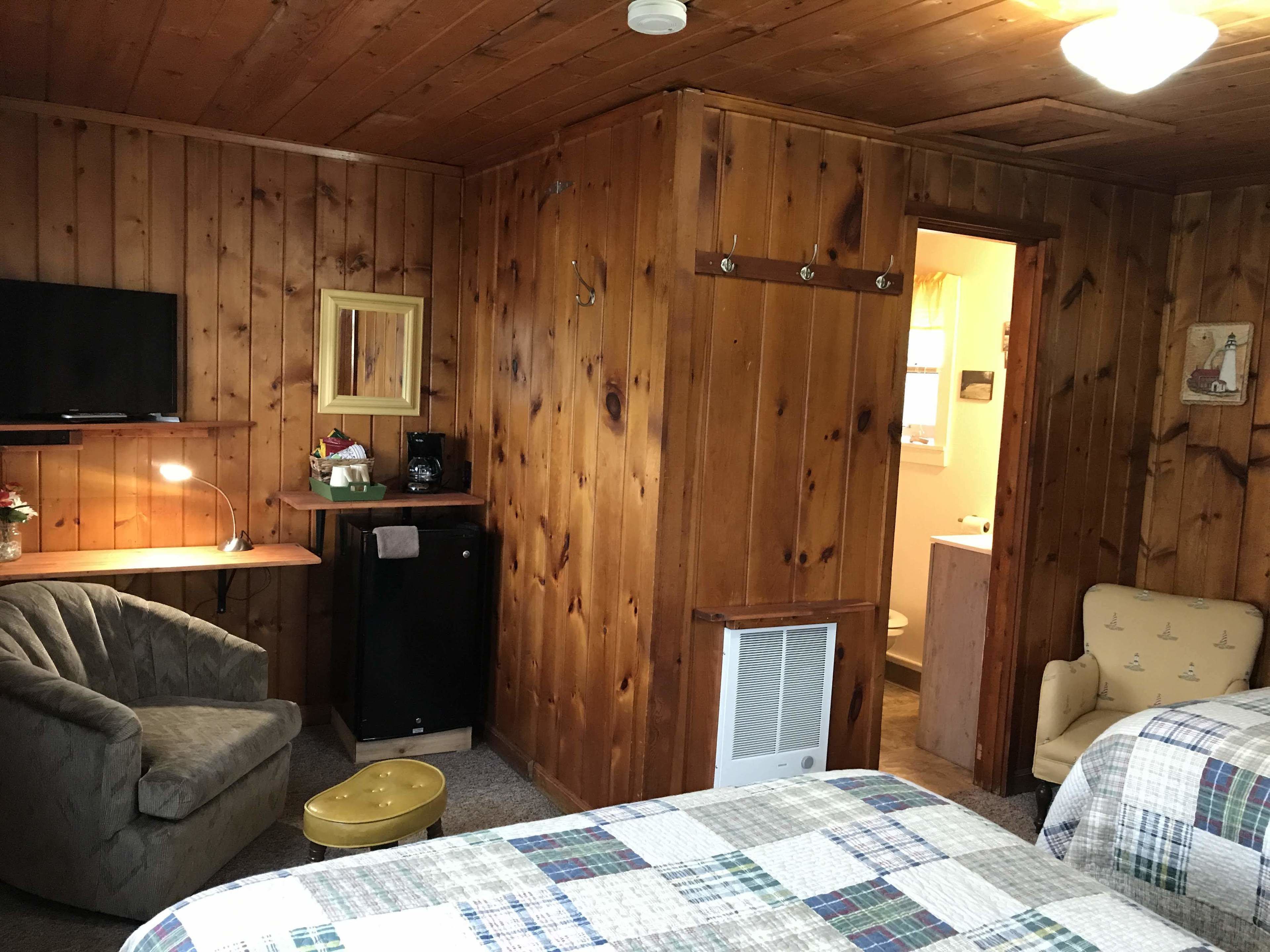 A cozy room with wooden walls, a television, a desk with a lamp, a refrigerator, a bed with a quilt, and a chair.