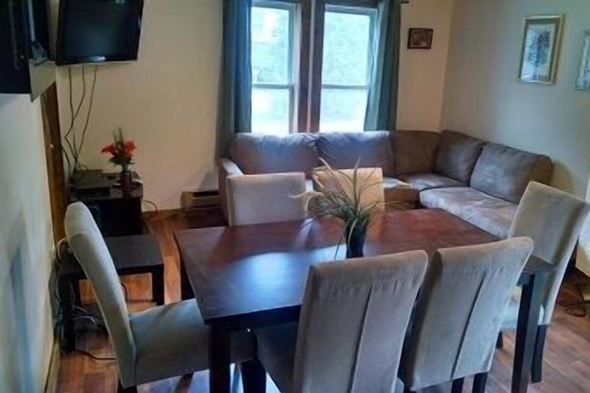 A cozy living room with a dark wooden dining table surrounded by chairs, a sofa, and a TV mounted on the wall.