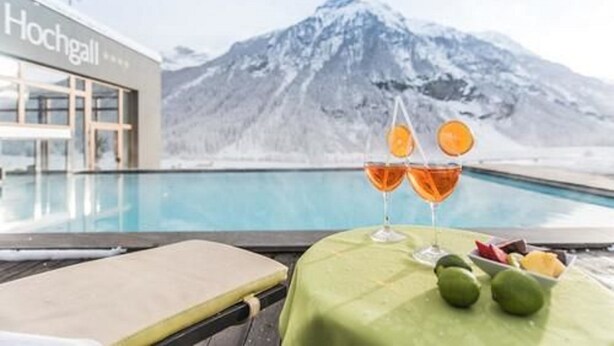 Vista de la piscina con montañas al fondo, una mesa con frutas y bebidas, y un edificio con el rótulo "Hochgall".