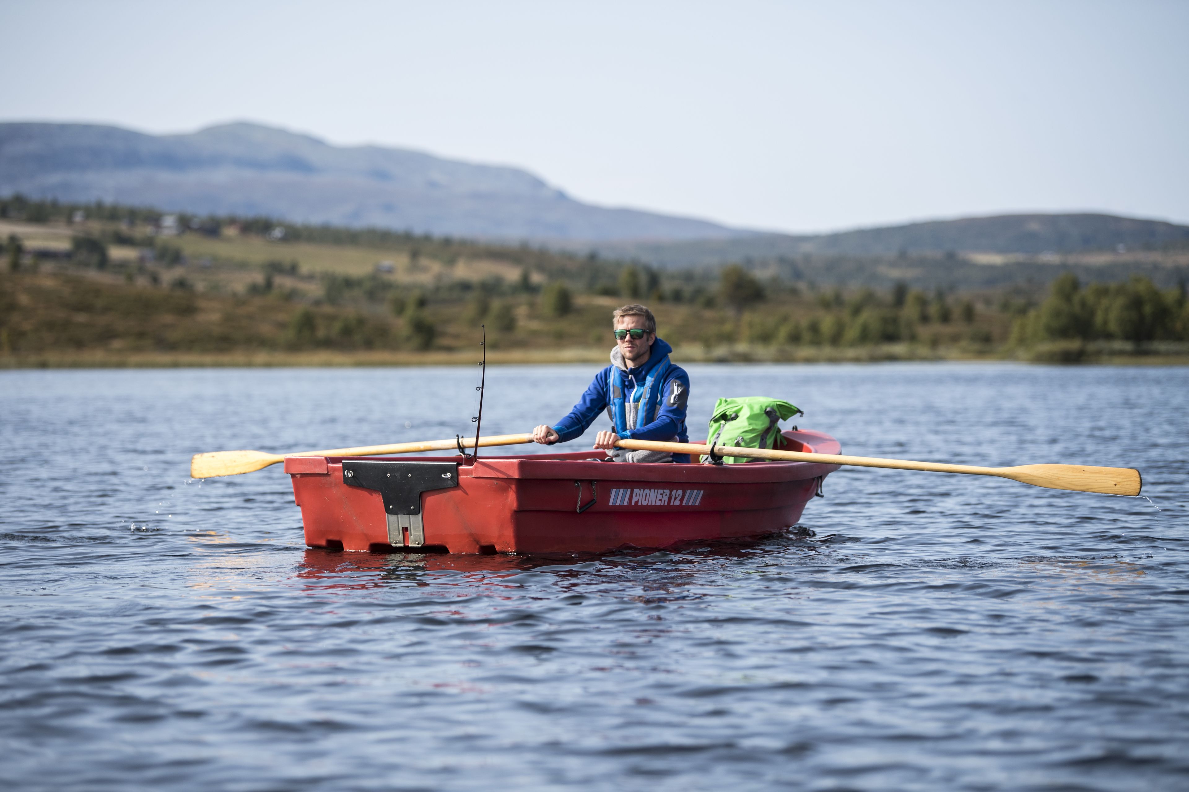 Una persona en un bote rojo con una caña de pescar, en un lago tranquilo con montañas al fondo.
